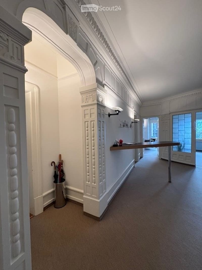Elegant hallway with ornate white paneling, a small console table with a vase, and a coat stand with umbrellas.