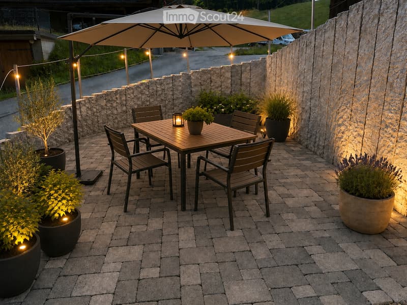 An outdoor patio area with a wooden table, four chairs, and a large umbrella, surrounded by potted plants and illuminated by string lights.