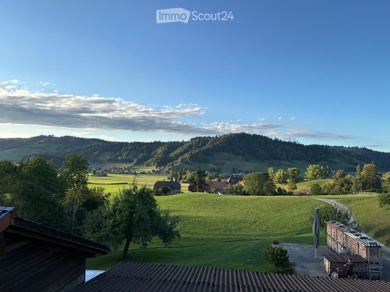 Panoramic view of a lush green valley with rolling hills, scattered houses, and a clear blue sky. The foreground shows a wooden structure and a stone wall, with a pathway leading into the landscape.