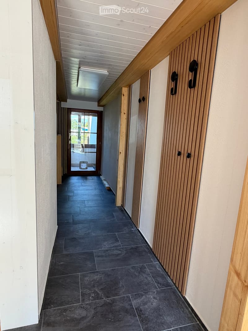 Modern hallway with dark tiled floor, white walls, wooden handrail, and a door leading to an outdoor area.
