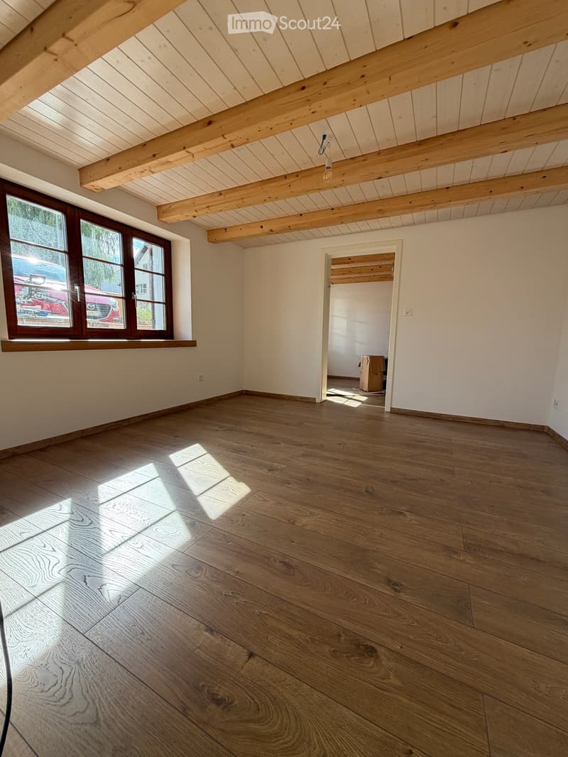 Empty room with wooden floor, exposed wooden beams on the ceiling, and a large window with wooden frame. Sunlight streams in, casting shadows on the floor.