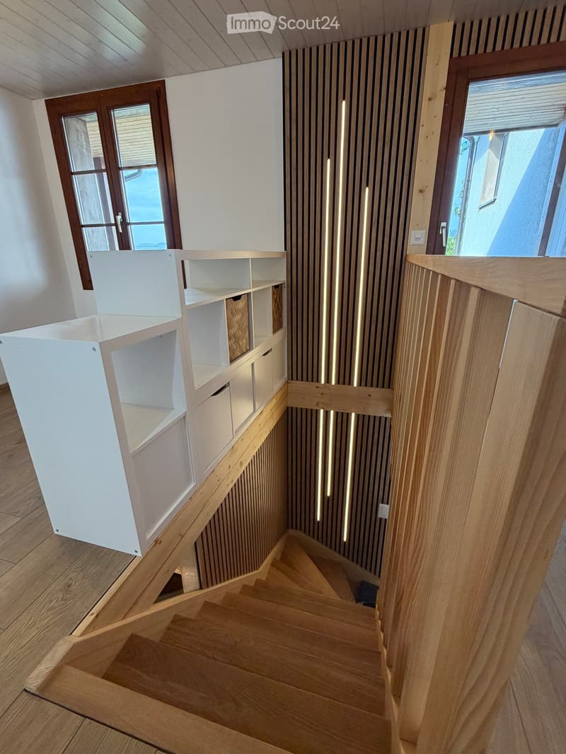 Modern wooden staircase with white built-in shelving unit, vertical light strips on wall, and large windows providing natural light.