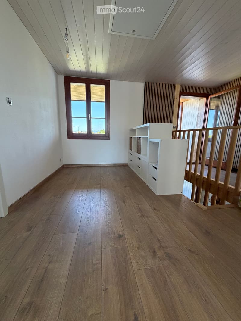 Empty room with wooden flooring, white walls, and a window with blinds. A white shelving unit is positioned near the staircase railing.