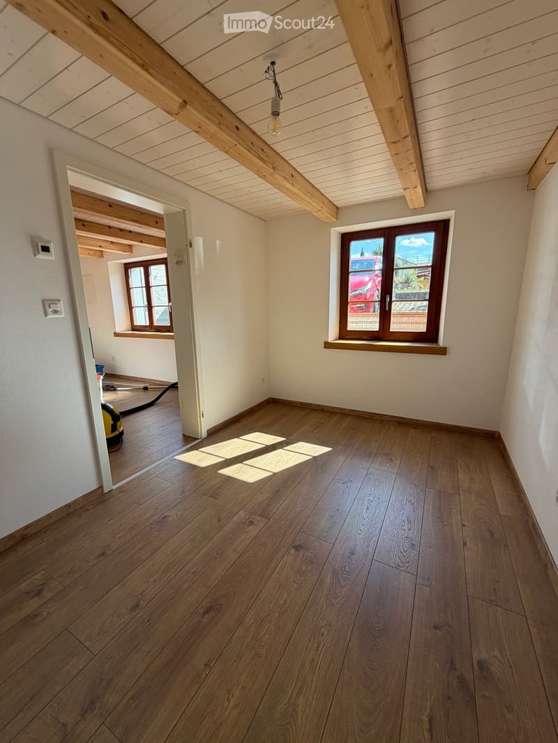 Empty room with wooden flooring, white walls, exposed wooden beams on the ceiling, and large windows with wooden frames. Sunlight streams in, illuminating the space.