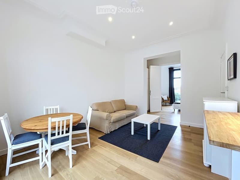 Bright, modern living and dining area with light-colored furniture, including a sofa, coffee table, dining table with chairs, and a kitchen counter visible in the background.