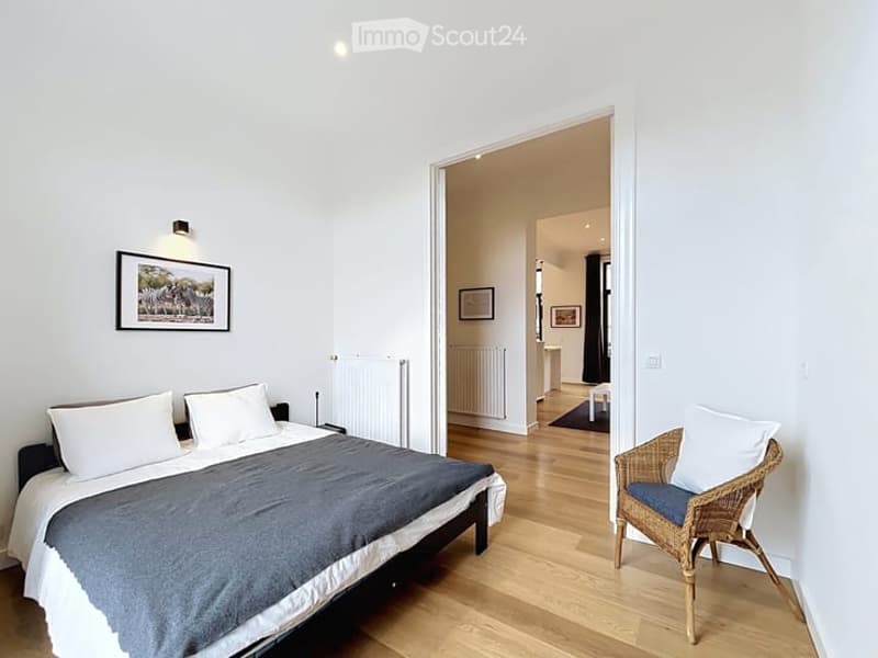 Cozy bedroom with a double bed, wicker chair, and picture frame on the wall. The room has light wooden flooring and a view into an adjacent bathroom.