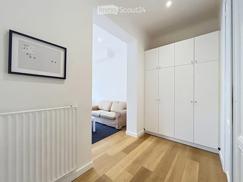 Modern hallway leading to a living area with a light-colored sofa, coffee table, and built-in white wardrobes. The hallway features a radiator and a framed picture on the wall.