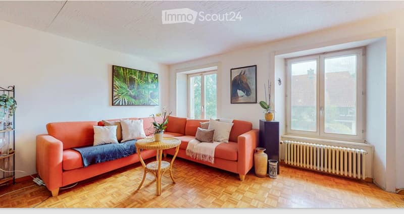 L-shaped orange sofa, wooden coffee table, white walls, wooden floor, radiator, large windows, paintings, plants