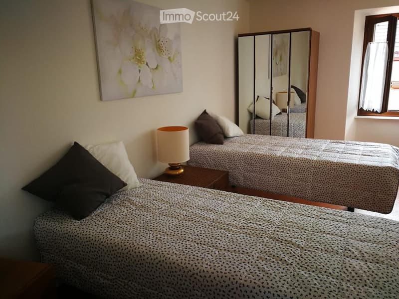 Cozy bedroom with two single beds, nightstands, lamps, and a large mirror. The room features a painting on the wall and a window with curtains.