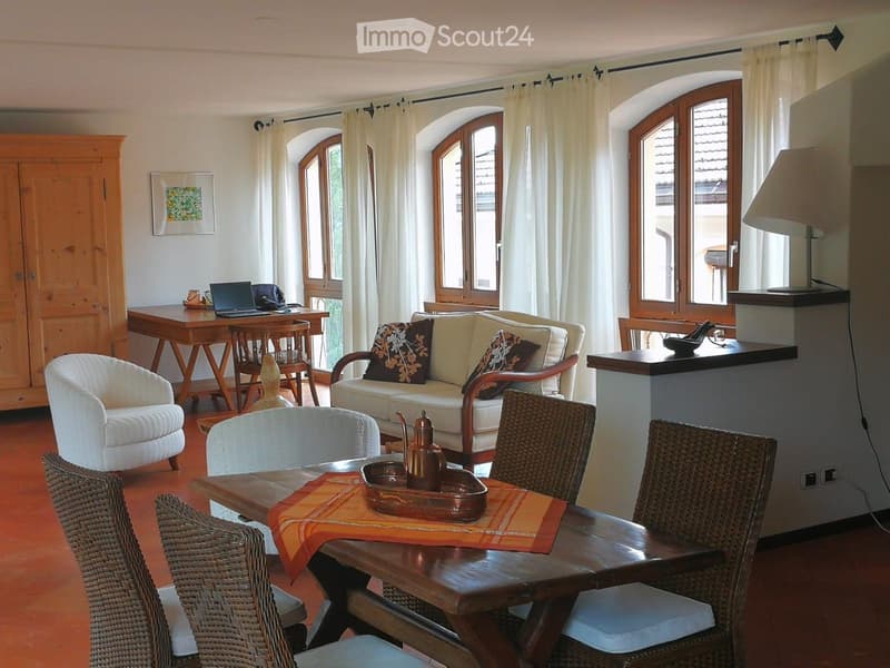 Bright living room with large arched windows, cream-colored sofa with patterned cushions, wooden coffee table with teapot, wicker chairs, white armchair, wooden dining table with laptop, and a wooden cabinet with a picture frame.