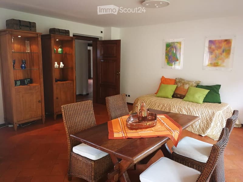 A cozy living space with a sofa, a dining table with chairs, a wooden cabinet, and decorative shelves. The room features a warm color palette with colorful cushions and artwork on the walls.