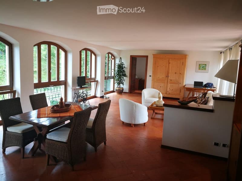 A well-lit living and dining area with wooden floors, large arched windows, and a balcony view. The space includes a dining table with chairs, a sofa, a laptop on a side table, and a potted plant.
