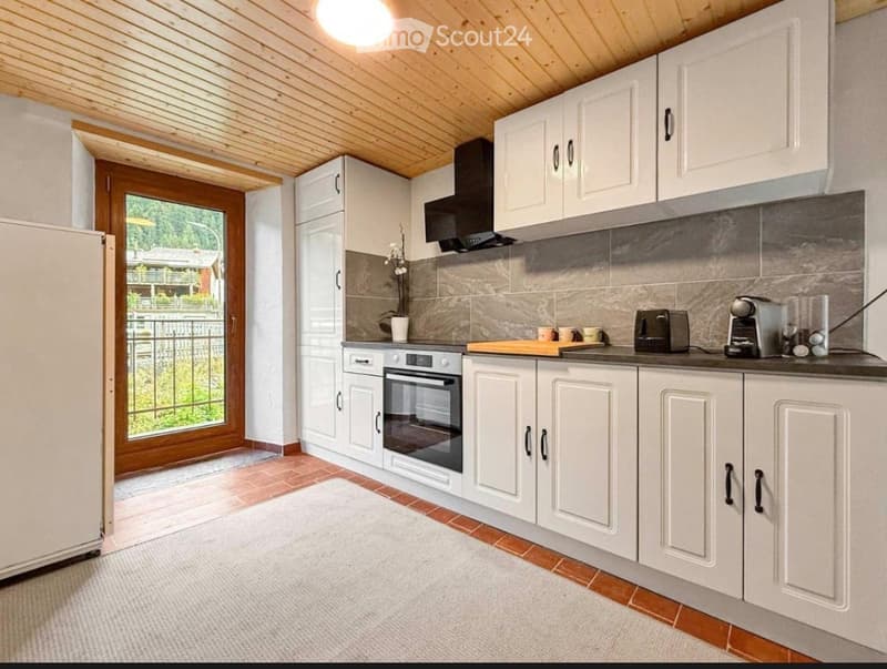 Modern kitchen with white cabinets, stainless steel appliances, and a view through a large window. Includes a refrigerator, oven, toaster, coffee maker, and a small flower vase on the counter.