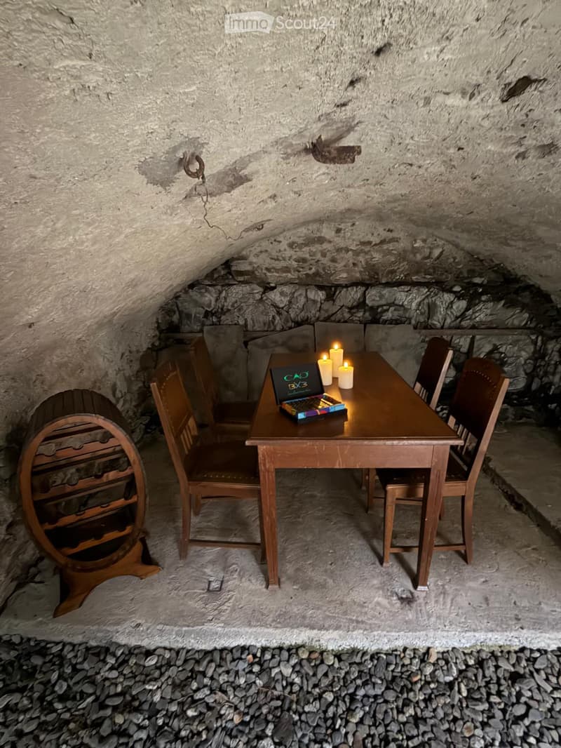 A rustic underground room with a wooden table, chairs, a laptop, candles, and a wine rack.