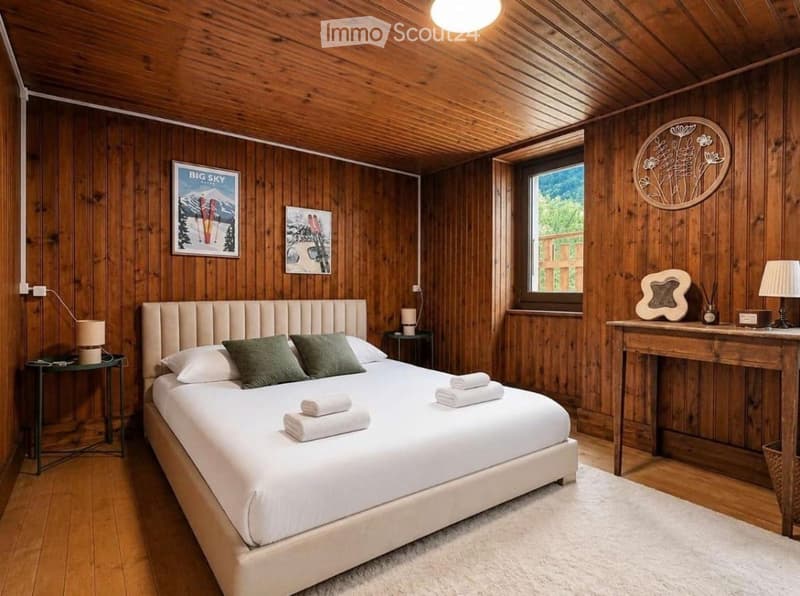 Cozy bedroom with wooden walls and ceiling, a large bed with white linens and green pillows, nightstands with lamps, a small table with a lamp, and decorative wall art.