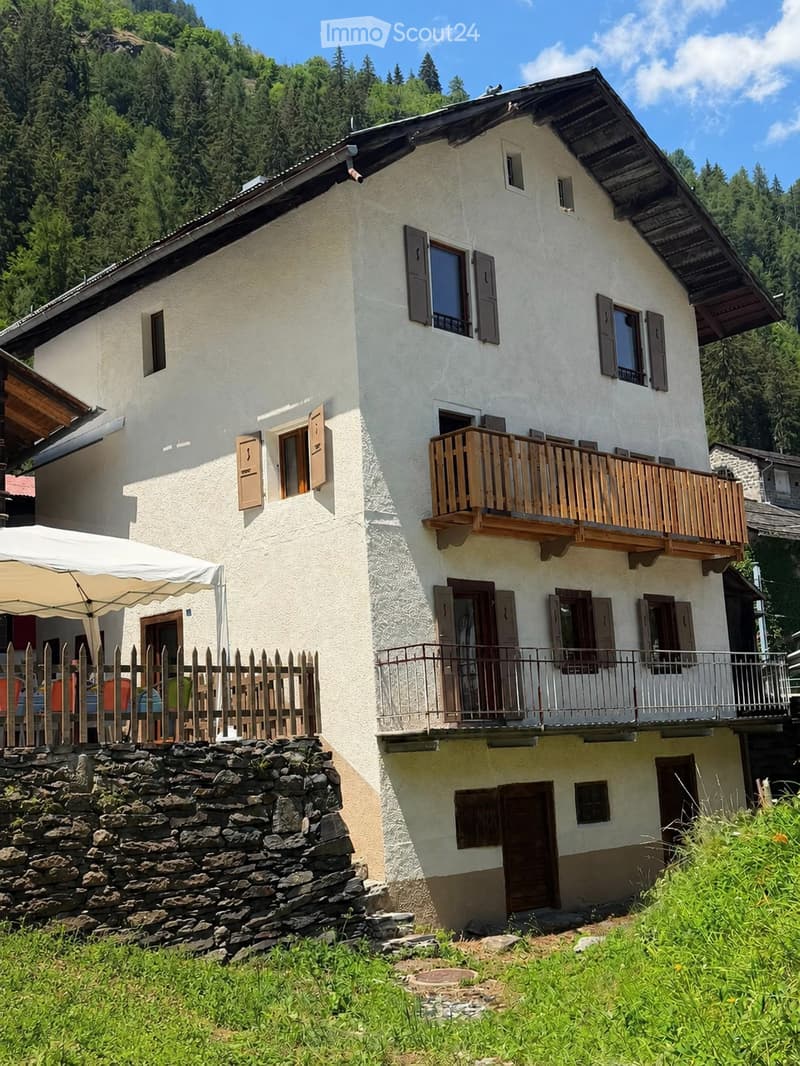 Two-story house with white walls, wooden shutters, and a wooden balcony. The house is surrounded by greenery and has a stone fence and patio area with outdoor furniture.