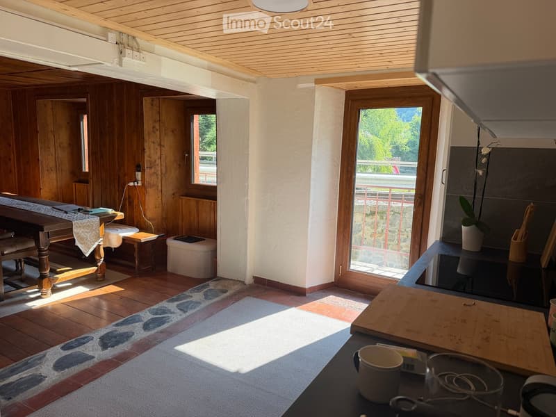 Bright, wooden-floored room with a dining table, chairs, a small stool, a cutting board, and a vase on a counter. Large windows offer a scenic view.