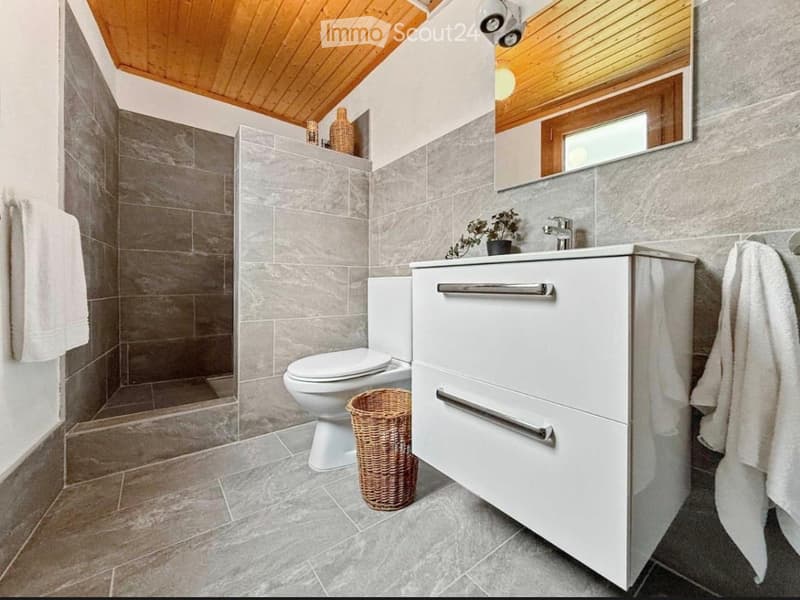 Modern bathroom with grey tiled walls and floor, white vanity with drawers, toilet, and a wicker basket on the floor.