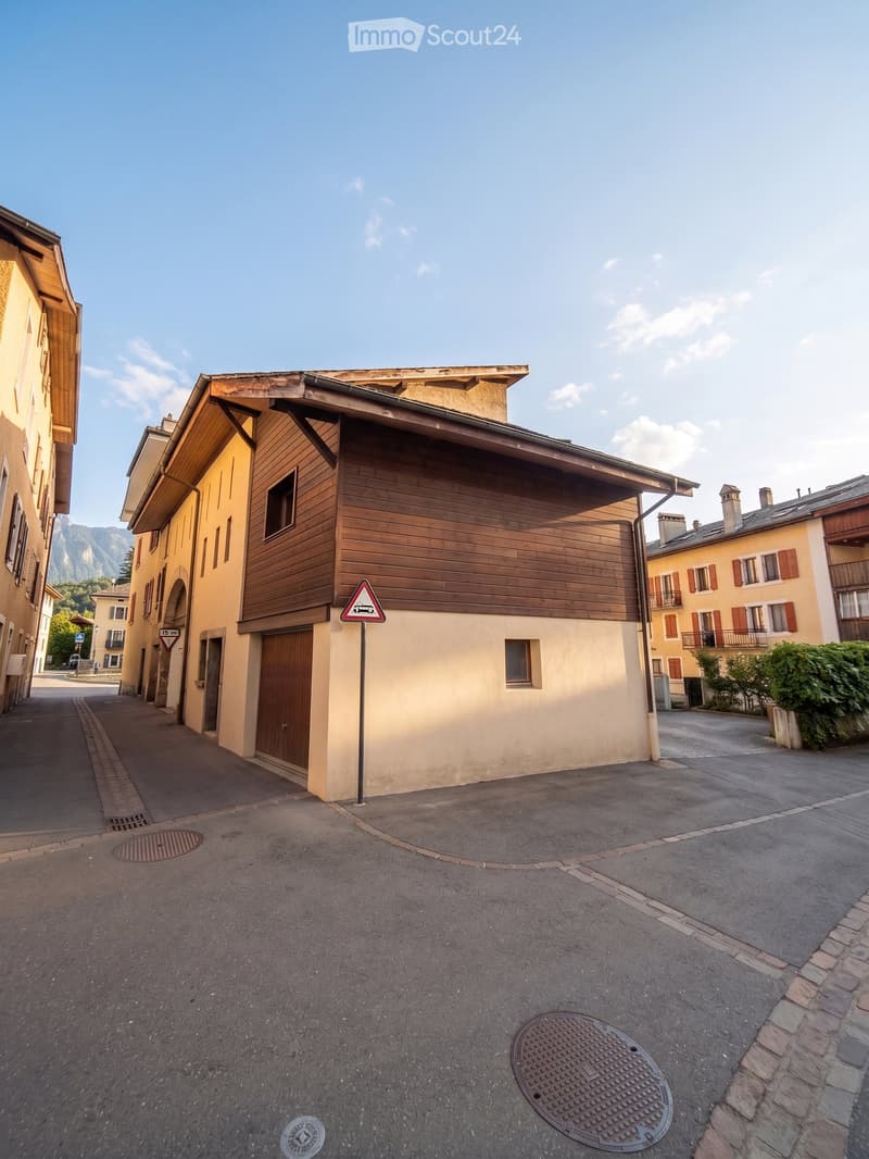 A multi-story building with wooden siding and a sloped roof, situated on a narrow street with a clear blue sky and distant mountains in the background.