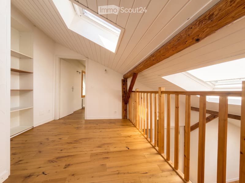Bright, airy hallway with wooden flooring, white walls, and exposed wooden beams. Features built-in shelving on one side and a staircase leading upwards with a wooden railing. Skylights provide natural light.