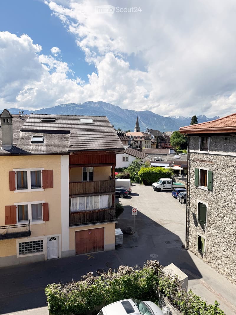 A multi-story residential building with a balcony, surrounded by other houses and parked cars, with a scenic mountain view in the background.