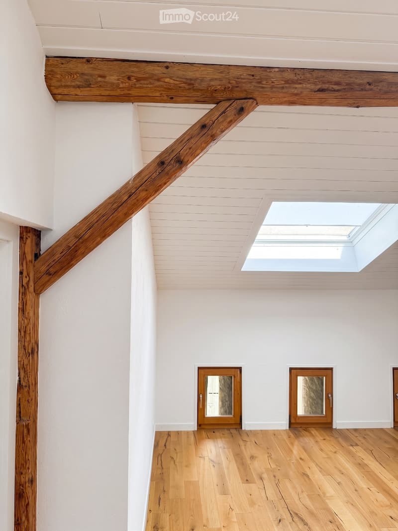 An empty room with wooden flooring, white walls, and a skylight window. The room features a wooden staircase railing and three wooden doors with glass panels.