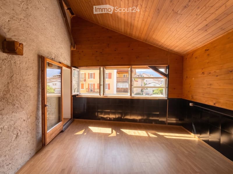 An empty room with wooden flooring, wooden ceiling, and large windows offering a view of the town and mountains.