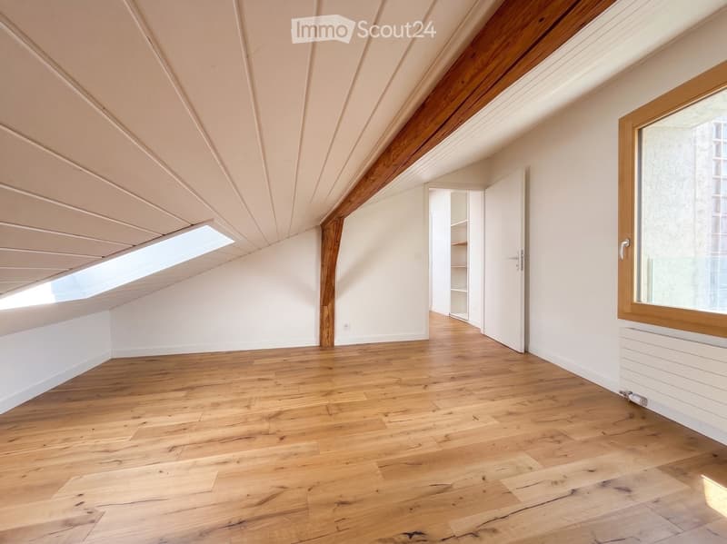 An empty attic room with wooden flooring, white walls, and exposed wooden beams. Features a large skylight and a window with wooden frame, along with a small open closet area.