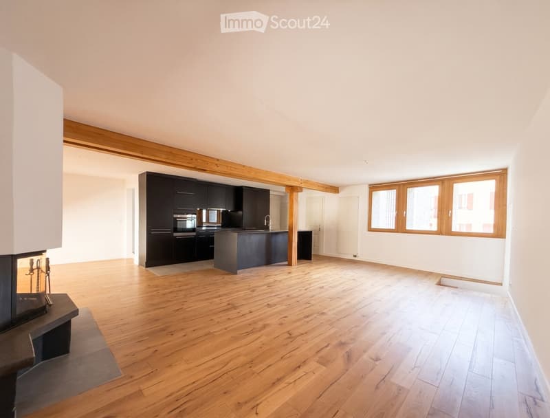 Open-plan living and kitchen area with wooden flooring, exposed wooden beams, modern black kitchen cabinets, central island, and a fireplace on the left side.
