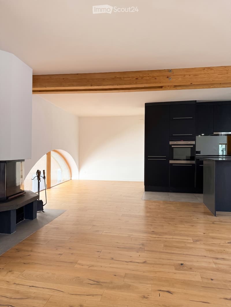 An empty room with wooden flooring, white walls, and a fireplace on the left side. The room features exposed wooden beams on the ceiling and a modern kitchen area with dark cabinets and stainless steel appliances on the right side.