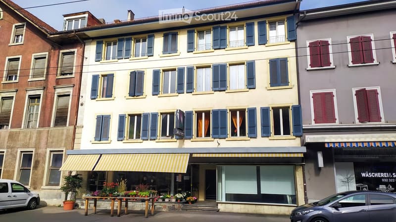 A multi-story building with blue shutters and a storefront on the ground floor, featuring a yellow striped awning and potted plants displayed outside.