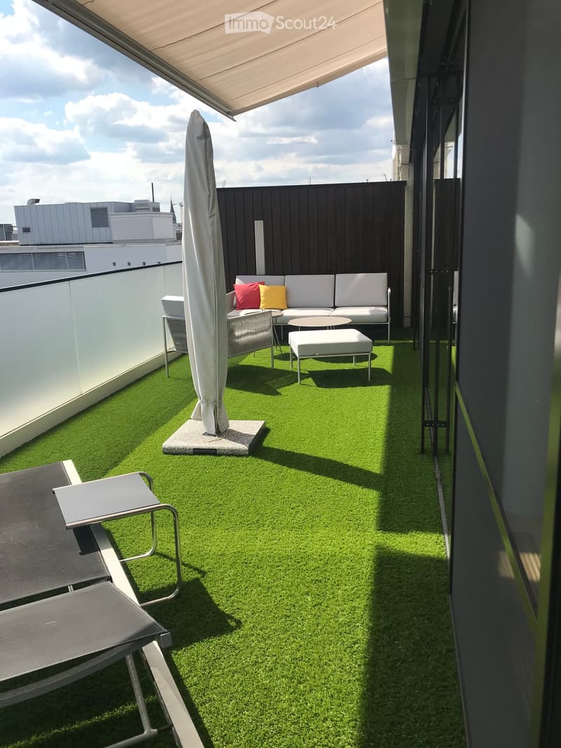 A modern rooftop terrace with artificial grass, a white sofa with colorful cushions, a small table, chairs, a stool, and a closed umbrella.