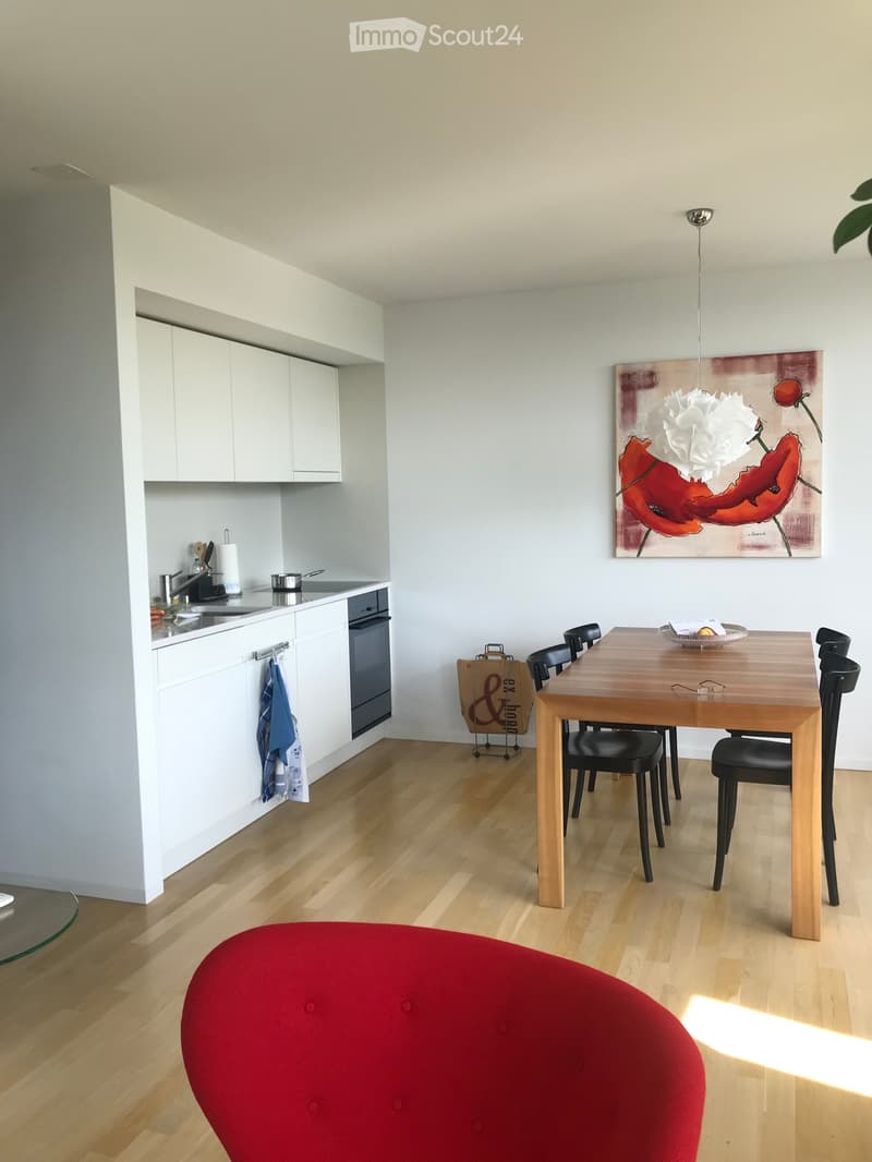 Modern open-plan kitchen and dining area with white cabinets, wooden dining table, black chairs, red round stool, and a large abstract painting on the wall.