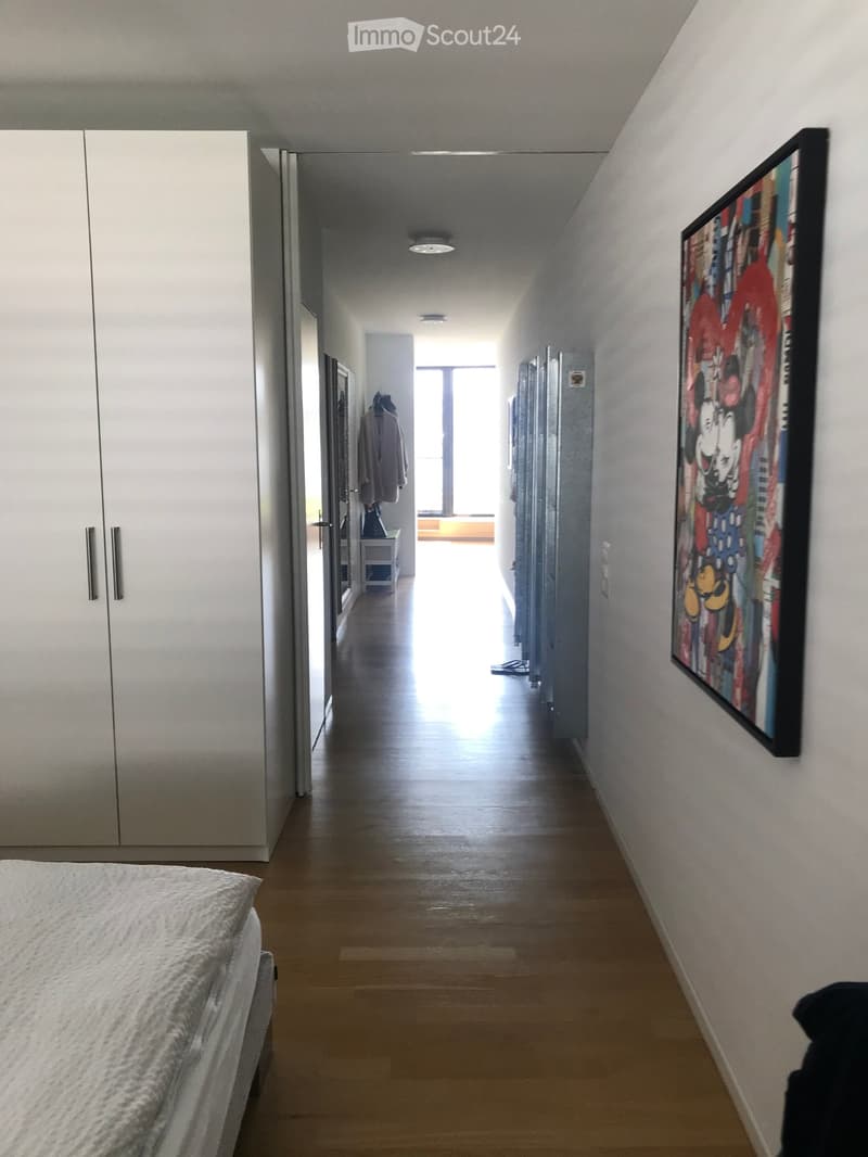 Modern hallway with wooden flooring, white walls, and a large painting on the right side. A bed is partially visible on the left, and a closet with mirrored doors is on the left wall. Natural light streams in from a window at the end of the hallway.