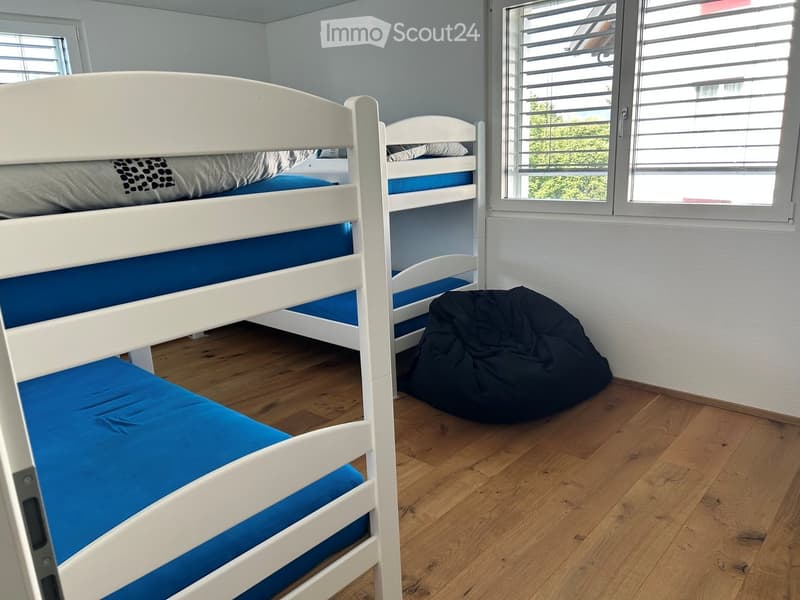 A small bedroom with two bunk beds and a bean bag chair, wooden flooring, and large windows with blinds.
