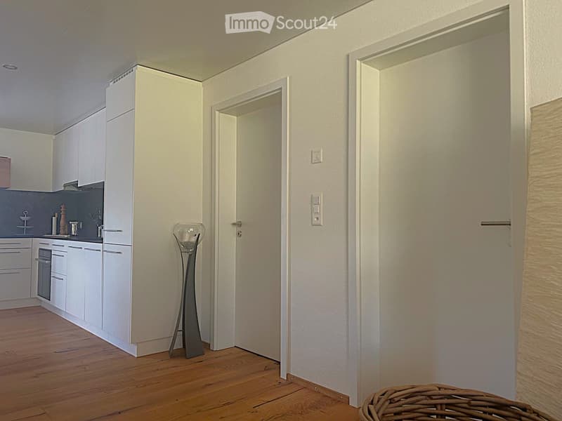 Modern, minimalist kitchen with white cabinets, a sink, and a decorative vase in the foreground. The space features wooden flooring and a sleek design.
