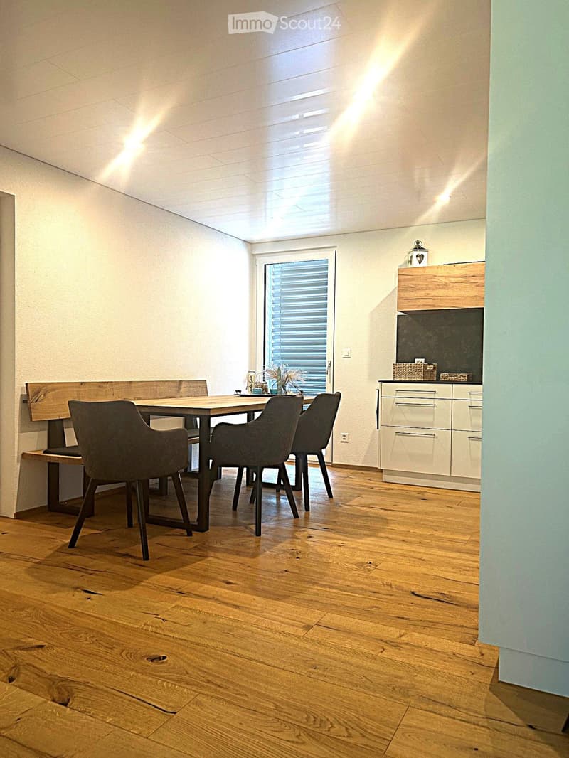 Bright dining area with wooden floor, modern dining table, four chairs, and a built-in bench along the wall. A small cabinet with drawers and a decorative item sits nearby.