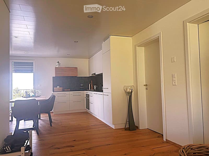 Modern kitchen and dining area with white cabinets, wooden flooring, and a large window overlooking a balcony. Includes a dining table with chairs, a floor lamp, and various kitchen appliances.