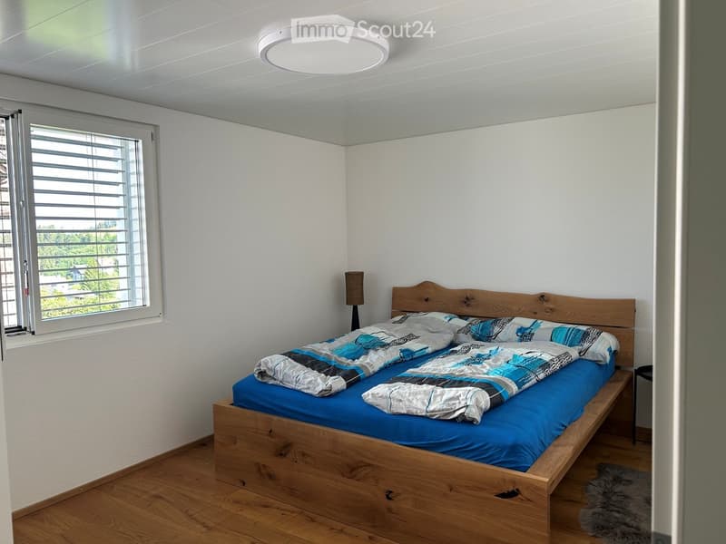 A modern bedroom with a wooden bed, blue bedding, a table lamp, and a window with blinds.