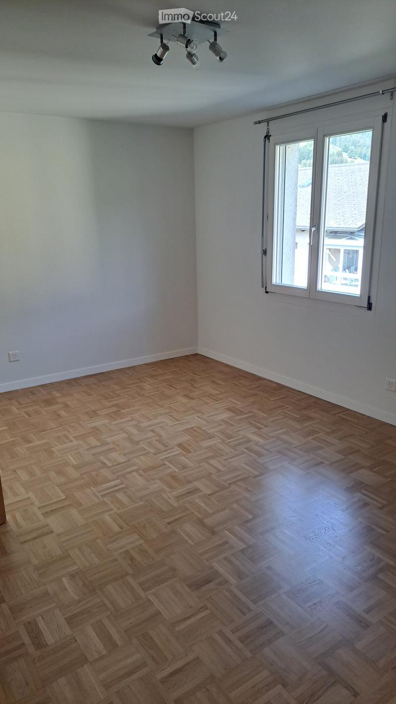 Empty room with herringbone-patterned wooden flooring, white walls, large window with blinds, and recessed ceiling lighting.