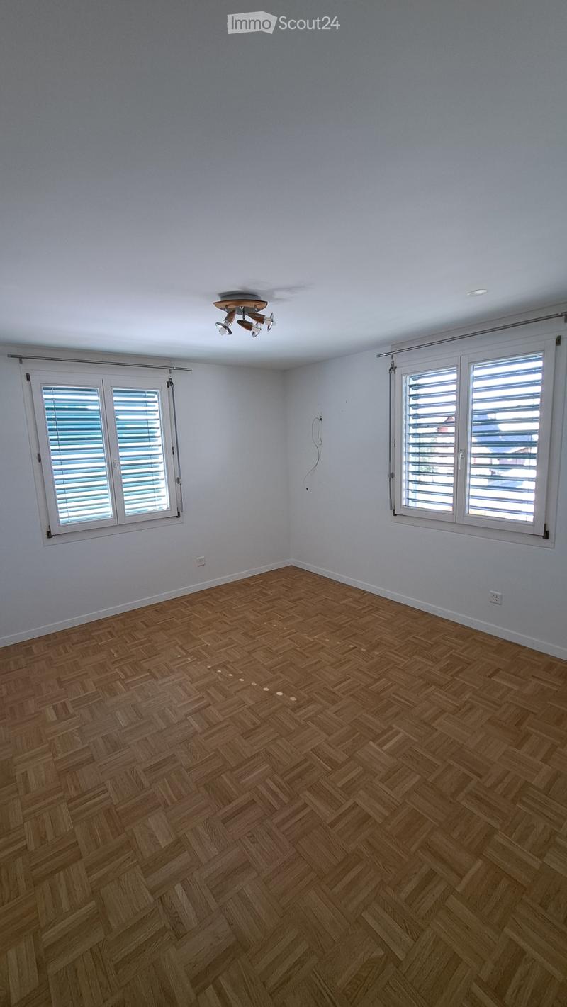 Empty room with wooden herringbone flooring, white walls, and large windows with blinds. Ceiling light fixture visible.