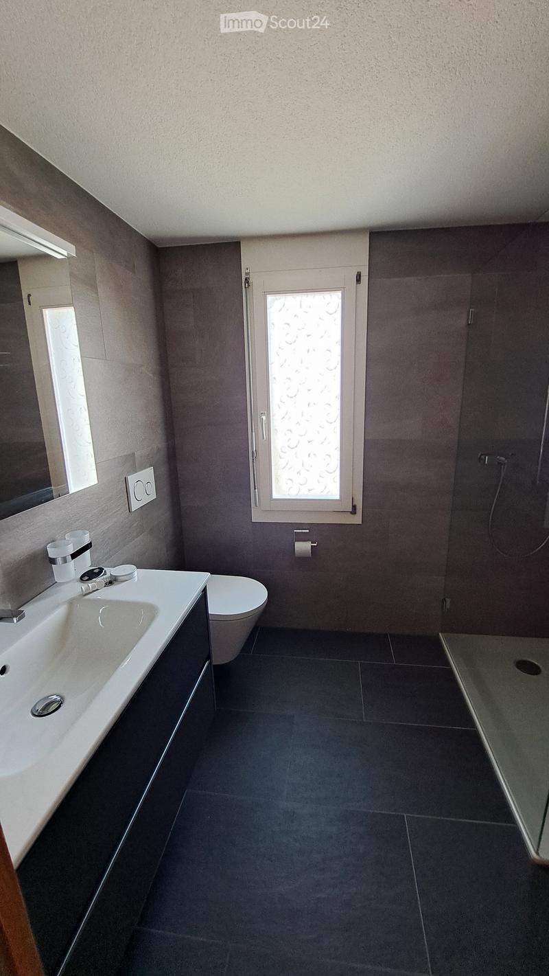 Modern bathroom with dark tiled floor, white sink, toilet, and a glass shower enclosure. Features a decorative window with frosted glass and a mirror above the sink.
