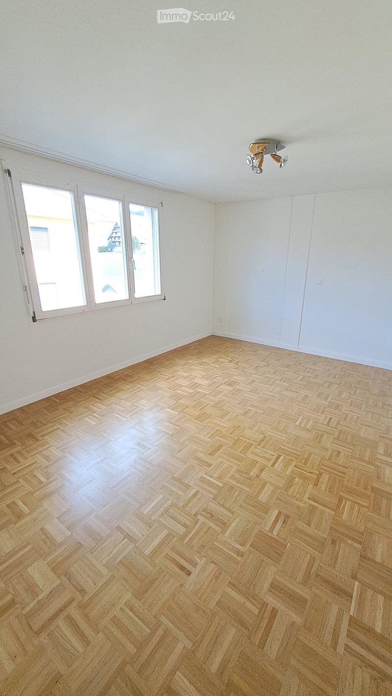 Empty room with light-colored walls, wooden herringbone flooring, large windows, and a ceiling light fixture.