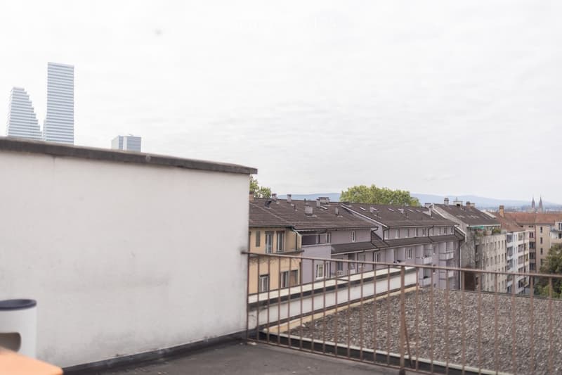 Roof terrace with metal railing, overlooking a cityscape with residential buildings, trees, and distant skyscrapers.