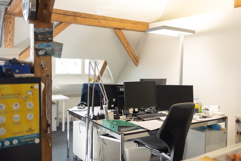 Office space with a desk, computer setup, chair, filing cabinet, and various office supplies. Exposed wooden beams and slanted ceiling.