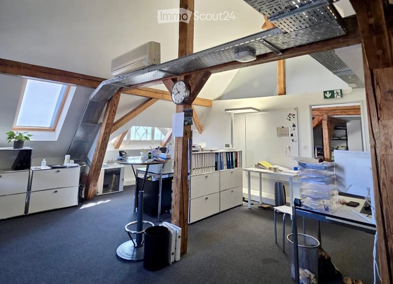 Office space with exposed wooden beams, skylights, and modern furniture including tables, chairs, filing cabinets, and a laptop. The room features a clock on the wall and a potted plant on a cabinet.
