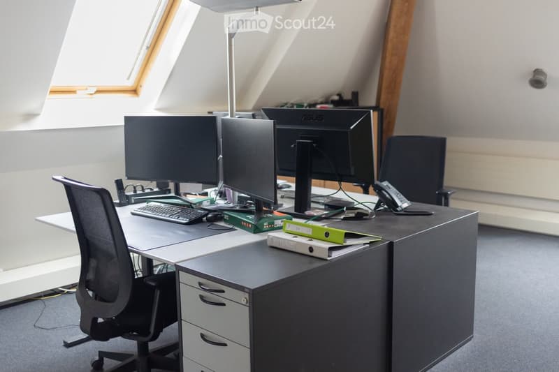 Modern office space with a large desk, multiple computer monitors, ergonomic chair, filing cabinet, and a floor lamp near a skylight window.