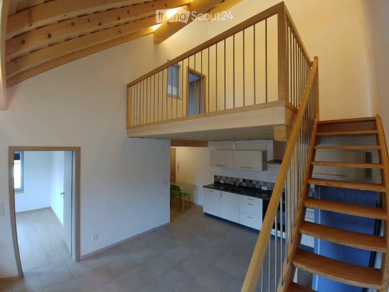 Open-plan hallway with wooden staircase leading to an upper level, kitchen area visible with cabinets, countertop, and a green chair, tiled floor, and white walls.