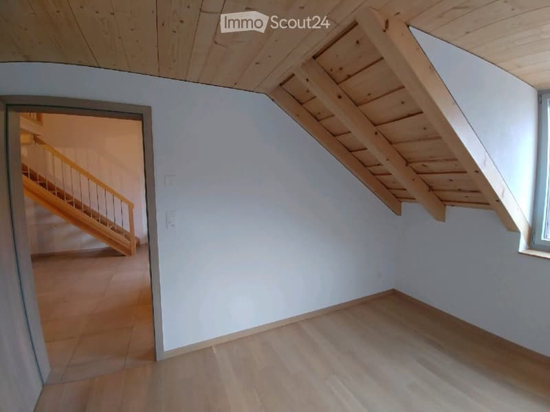 Empty room with light wooden flooring, white walls, and a wooden staircase leading to an attic space with exposed wooden beams.