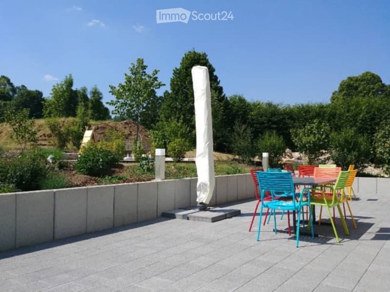 Outdoor patio with colorful chairs and a table, surrounded by greenery and a low concrete wall.